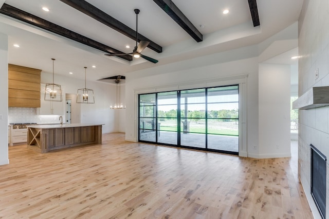 unfurnished living room with a tile fireplace, ceiling fan with notable chandelier, beam ceiling, light hardwood / wood-style flooring, and sink