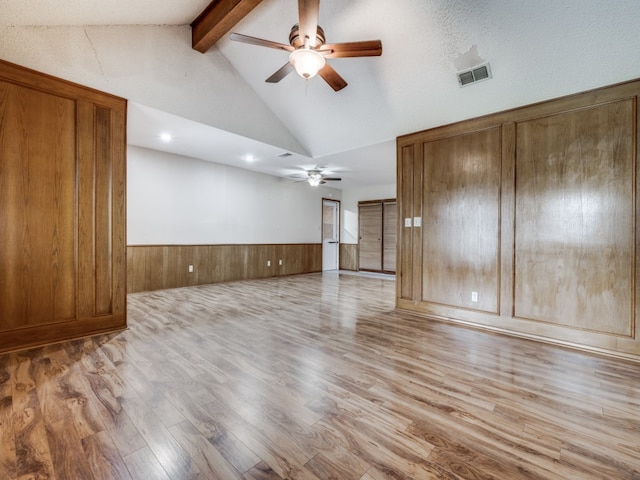 unfurnished living room with wood walls, ceiling fan, light wood-type flooring, and vaulted ceiling with beams