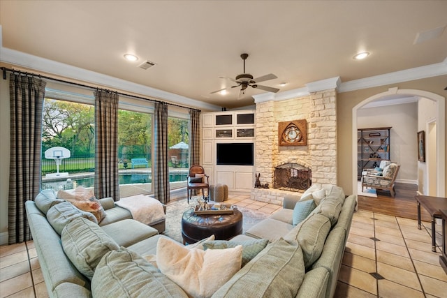 living room featuring light tile flooring, ceiling fan, a fireplace, crown molding, and built in shelves