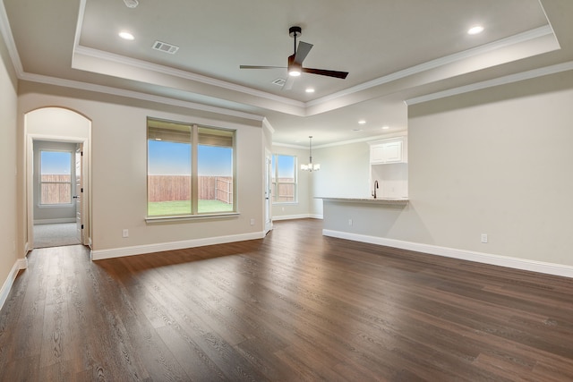 unfurnished living room with ceiling fan with notable chandelier, a tray ceiling, crown molding, and dark wood-type flooring