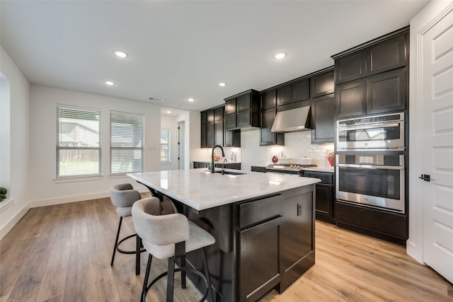 kitchen with stainless steel appliances, sink, light hardwood / wood-style flooring, a kitchen island with sink, and wall chimney exhaust hood