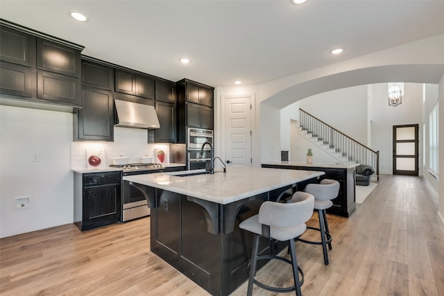 kitchen featuring an inviting chandelier, stainless steel appliances, light hardwood / wood-style floors, a kitchen island with sink, and wall chimney exhaust hood