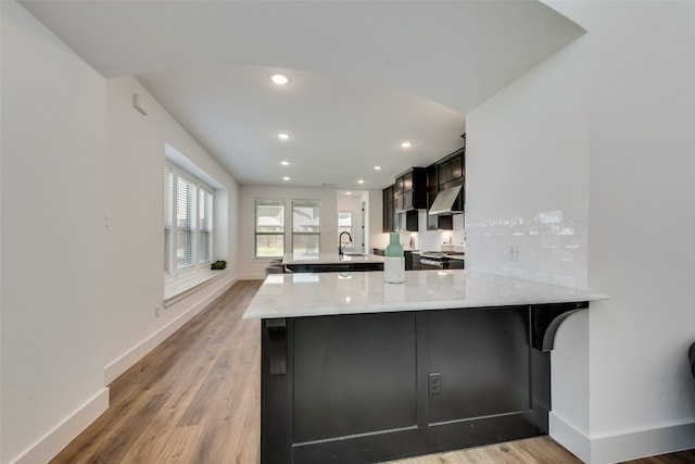 kitchen with light stone countertops, kitchen peninsula, wall chimney range hood, and wood-type flooring