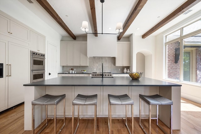 kitchen featuring white cabinets, beamed ceiling, a center island with sink, stainless steel double oven, and light wood-type flooring