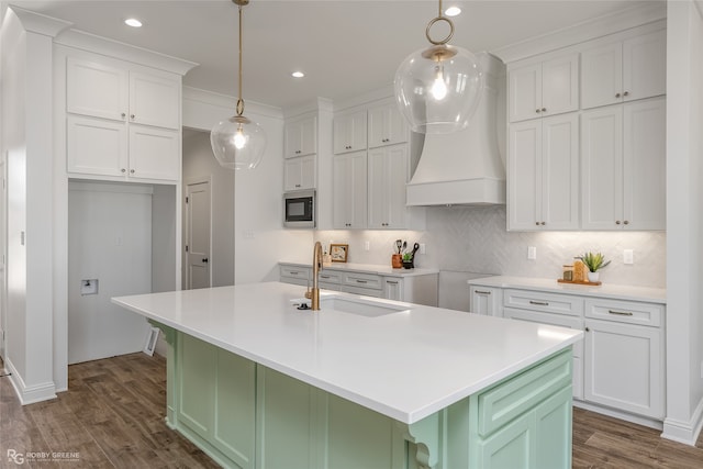 kitchen with sink, stainless steel microwave, a kitchen island with sink, white cabinetry, and custom range hood