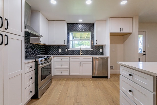 kitchen with appliances with stainless steel finishes, light hardwood / wood-style floors, white cabinetry, wall chimney exhaust hood, and backsplash