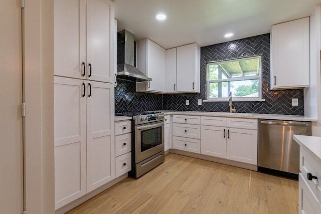 kitchen with appliances with stainless steel finishes, white cabinetry, light hardwood / wood-style flooring, wall chimney range hood, and sink