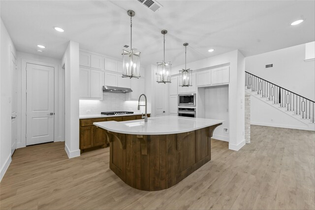 kitchen featuring appliances with stainless steel finishes, white cabinets, a center island with sink, decorative light fixtures, and light wood-type flooring