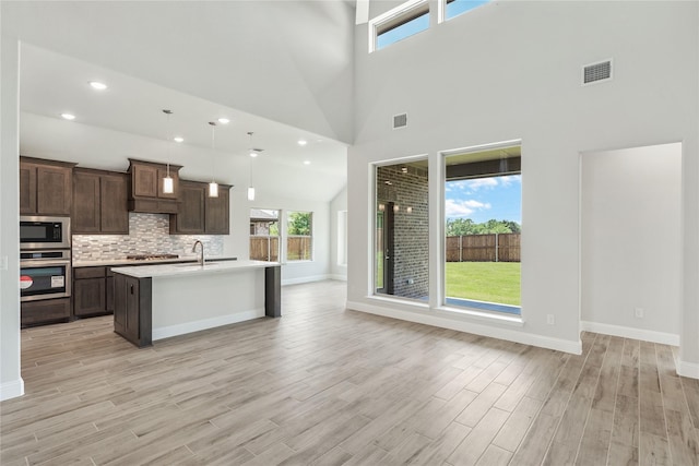 kitchen with stainless steel appliances, a high ceiling, pendant lighting, a kitchen island with sink, and light wood-type flooring