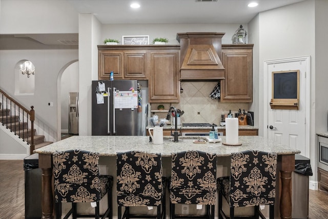 kitchen featuring stainless steel refrigerator, light stone countertops, dark hardwood / wood-style flooring, a kitchen island with sink, and custom range hood