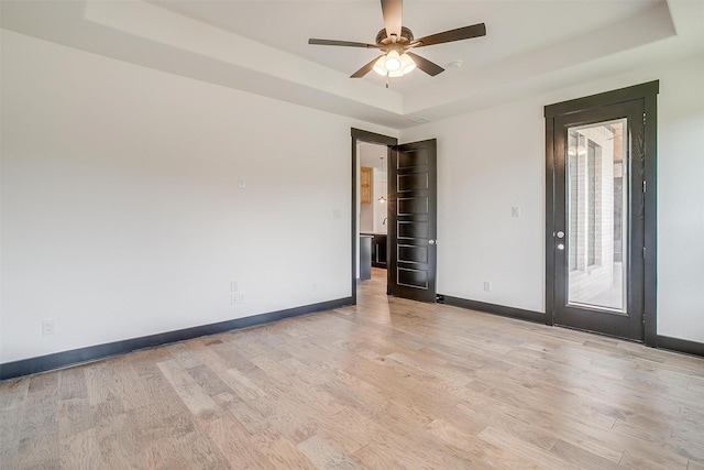 empty room featuring ceiling fan, light wood-type flooring, a raised ceiling, and baseboards