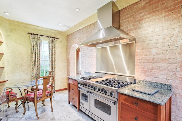 kitchen with wall chimney exhaust hood, light stone countertops, double oven range, light tile patterned floors, and brick wall