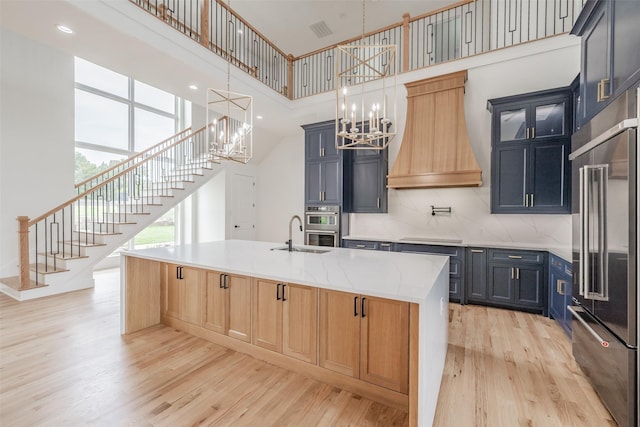 kitchen with appliances with stainless steel finishes, light wood-type flooring, a towering ceiling, custom exhaust hood, and a center island with sink