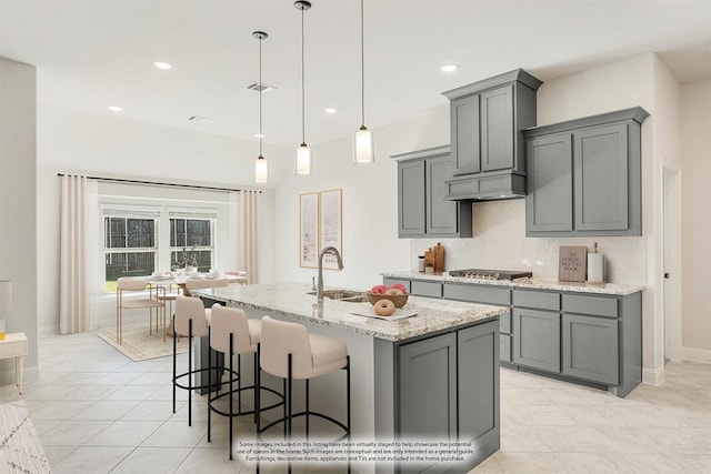 kitchen featuring light stone countertops, stainless steel gas stovetop, a center island with sink, gray cabinets, and hanging light fixtures