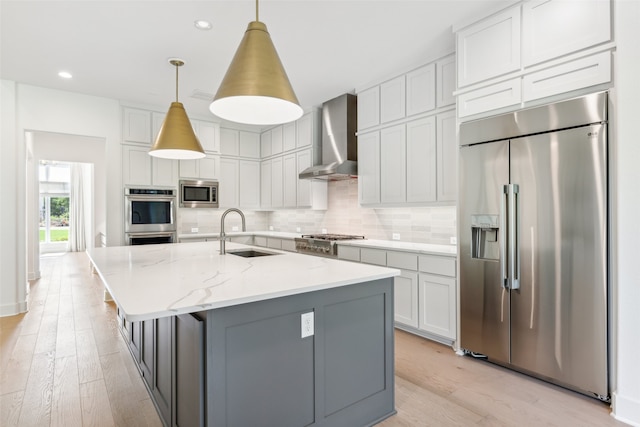 kitchen featuring wall chimney range hood, sink, built in appliances, light stone countertops, and an island with sink