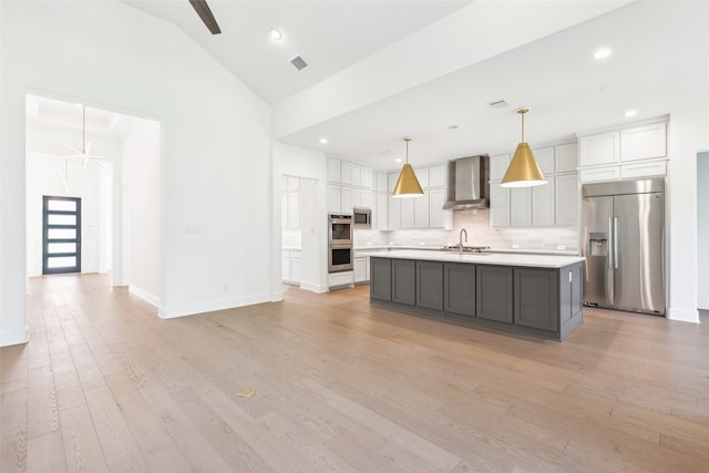 kitchen with wall chimney exhaust hood, pendant lighting, built in appliances, light hardwood / wood-style floors, and an island with sink