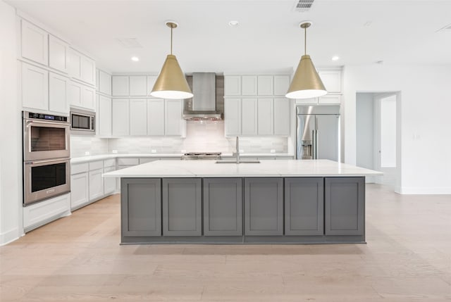 kitchen with built in appliances, wall chimney exhaust hood, an island with sink, and hanging light fixtures