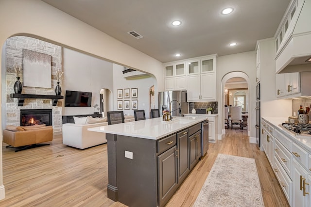 kitchen featuring appliances with stainless steel finishes, white cabinets, a stone fireplace, a kitchen island with sink, and decorative backsplash