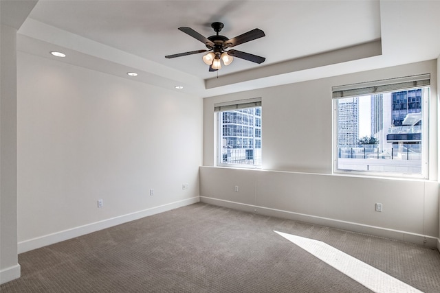 unfurnished room with a healthy amount of sunlight, dark colored carpet, ceiling fan, and a raised ceiling