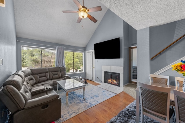 living room featuring high vaulted ceiling, light hardwood / wood-style flooring, a tiled fireplace, ceiling fan, and a textured ceiling