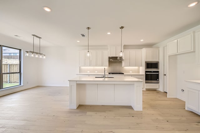 kitchen featuring light wood-type flooring, backsplash, a kitchen island with sink, stainless steel appliances, and decorative light fixtures