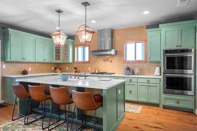 kitchen with light hardwood / wood-style flooring, stainless steel appliances, wall chimney exhaust hood, a center island with sink, and tasteful backsplash