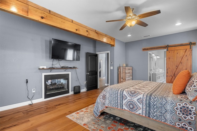 bedroom featuring a multi sided fireplace, wood-type flooring, ceiling fan, ensuite bathroom, and a barn door