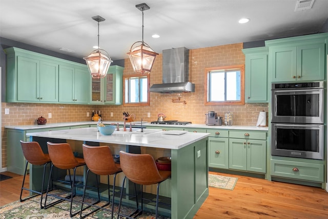 kitchen featuring appliances with stainless steel finishes, an island with sink, a kitchen breakfast bar, wall chimney exhaust hood, and light hardwood / wood-style flooring