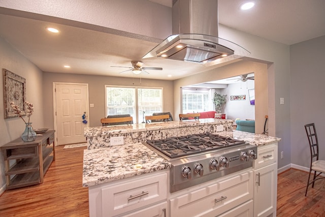 kitchen with ceiling fan, island range hood, wood-type flooring, stainless steel gas cooktop, and white cabinetry