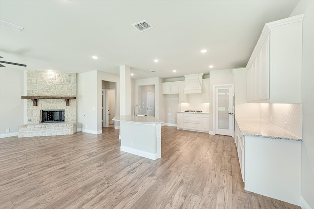 kitchen featuring light stone countertops, custom exhaust hood, an island with sink, white cabinetry, and a stone fireplace