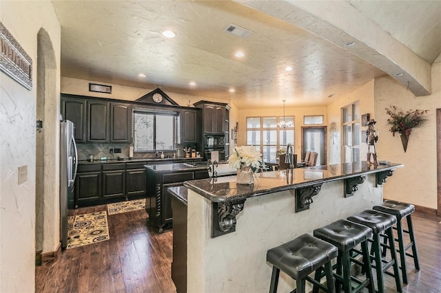 kitchen featuring a kitchen island with sink, a breakfast bar, decorative light fixtures, dark wood-type flooring, and dark stone countertops