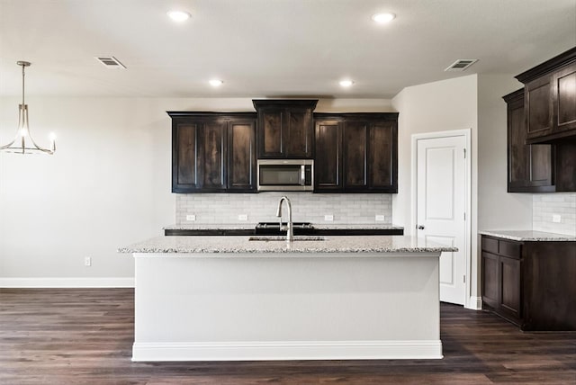 kitchen with light stone counters, dark hardwood / wood-style floors, an island with sink, backsplash, and dark brown cabinetry