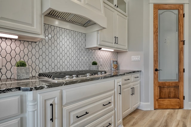 kitchen with white cabinets, backsplash, custom exhaust hood, stainless steel gas stovetop, and light hardwood / wood-style flooring