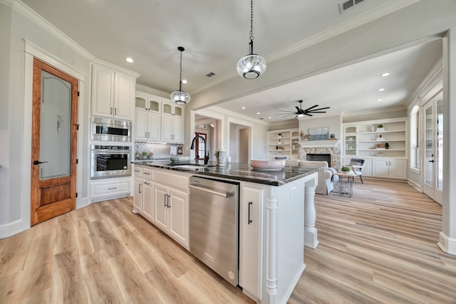 kitchen featuring appliances with stainless steel finishes, an island with sink, a stone fireplace, hanging light fixtures, and white cabinetry