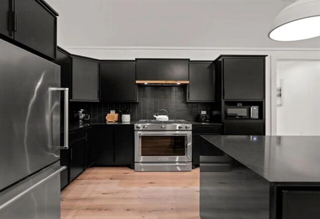 kitchen with light hardwood / wood-style flooring, appliances with stainless steel finishes, wall chimney range hood, tasteful backsplash, and dark stone counters