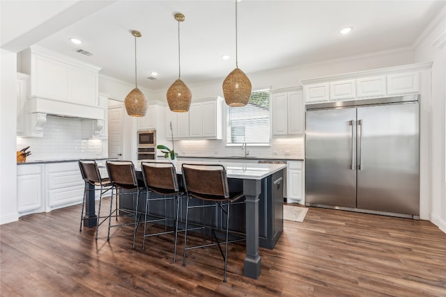 kitchen with built in appliances, decorative light fixtures, dark wood-type flooring, a center island with sink, and tasteful backsplash