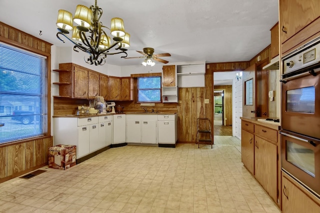 kitchen featuring stainless steel double oven, pendant lighting, ceiling fan with notable chandelier, and wood walls