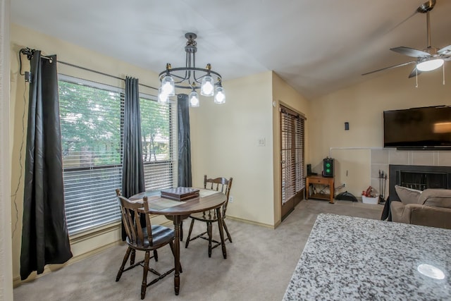 dining area with ceiling fan with notable chandelier, a tiled fireplace, light carpet, and vaulted ceiling