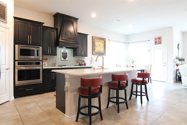 kitchen with appliances with stainless steel finishes, premium range hood, tasteful backsplash, an island with sink, and a breakfast bar