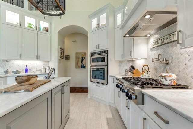 kitchen featuring arched walkways, baseboards, appliances with stainless steel finishes, premium range hood, and white cabinetry