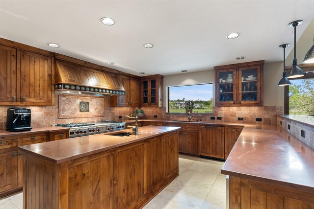 kitchen featuring hanging light fixtures, stainless steel stove, custom range hood, sink, and a center island with sink