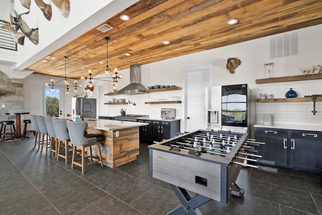 kitchen featuring wall chimney exhaust hood, a breakfast bar area, wooden ceiling, an island with sink, and pendant lighting