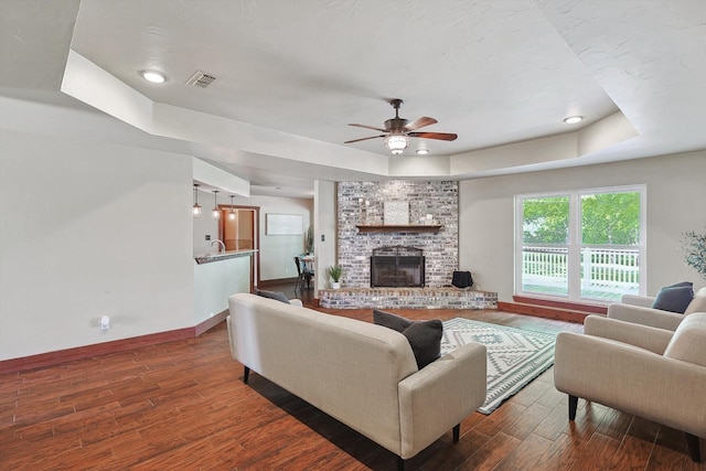 living room featuring a fireplace, ceiling fan, a tray ceiling, brick wall, and dark hardwood / wood-style floors