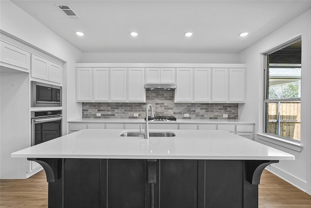 kitchen with appliances with stainless steel finishes, an island with sink, white cabinets, backsplash, and light hardwood / wood-style flooring