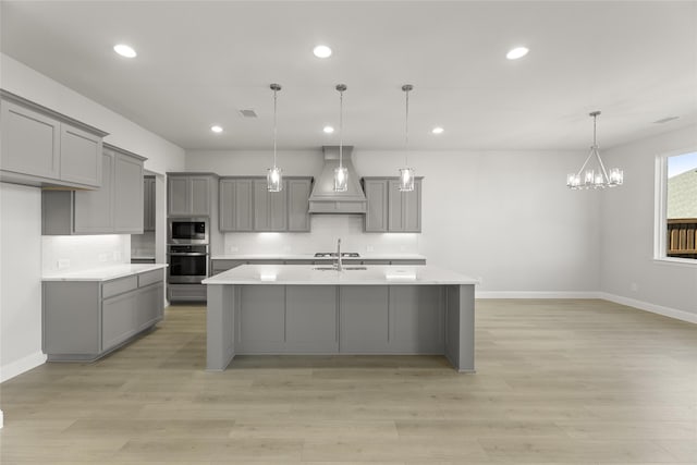 kitchen featuring stainless steel appliances, custom range hood, gray cabinets, pendant lighting, and a kitchen island with sink