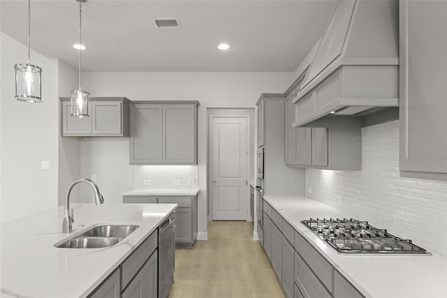 kitchen featuring sink, custom exhaust hood, light wood-type flooring, appliances with stainless steel finishes, and decorative light fixtures