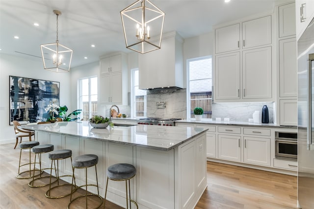 kitchen with pendant lighting, wall chimney exhaust hood, an island with sink, and white cabinets