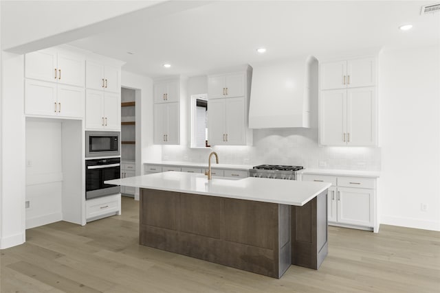 kitchen featuring a center island with sink, stainless steel microwave, wall chimney range hood, white cabinets, and wall oven