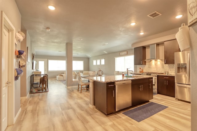 kitchen with dark brown cabinets, light hardwood / wood-style flooring, stainless steel appliances, tasteful backsplash, and wall chimney exhaust hood