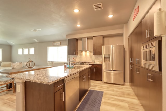 kitchen with appliances with stainless steel finishes, wall chimney exhaust hood, a kitchen island, backsplash, and light hardwood / wood-style flooring